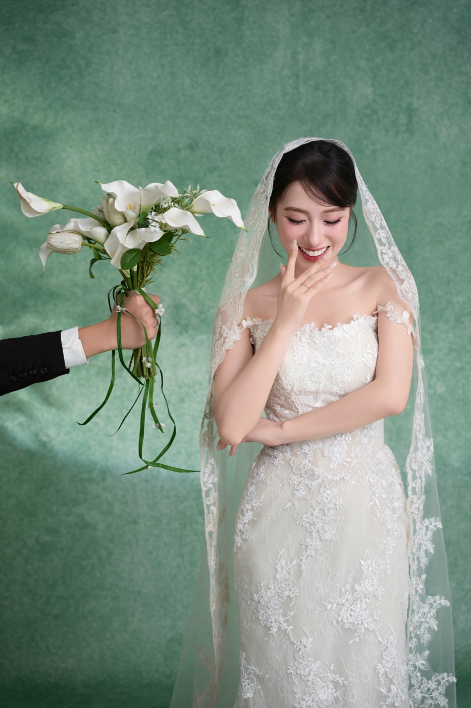 Cô dâu trong voan ren cúi đầu cười, một bàn tay chìa bó hoa từ bên trái