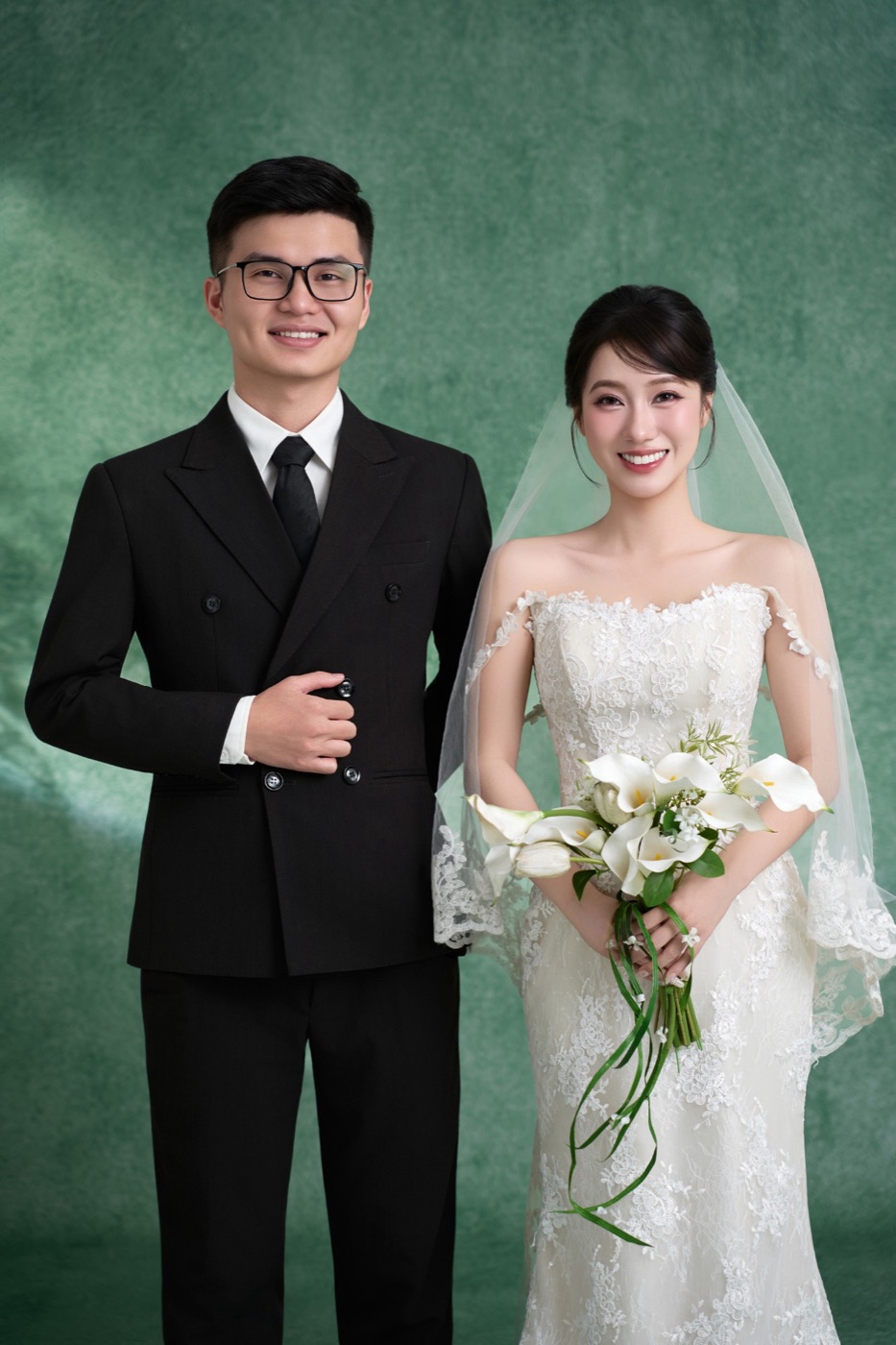 Đức Trịnh và Emi đứng cạnh nhau, chú rể vest đen, cô dâu cầm hoa rum